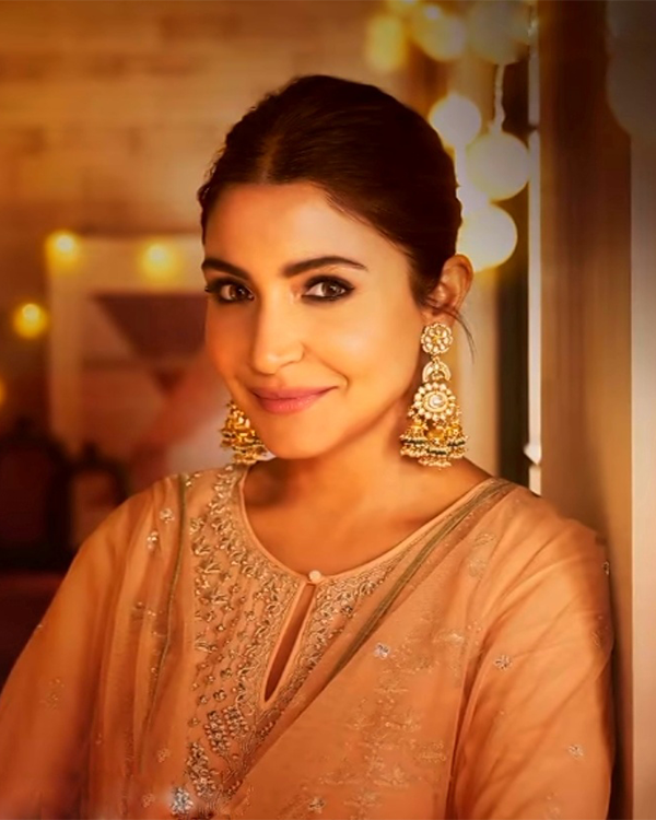 Anushka Sharma smiles wearing large gold jhumka earrings and a light peach embroidered outfit.
