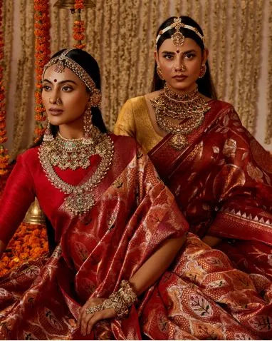 Two women in traditional red sarees with elaborate jewelry against a decorated background.