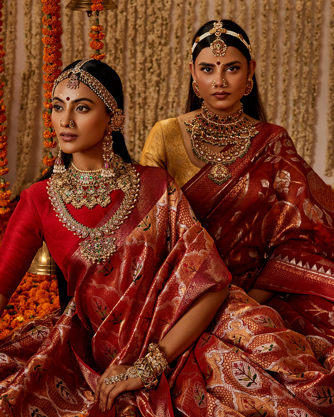 Two women in traditional red sarees with elaborate jewelry against a decorative background.