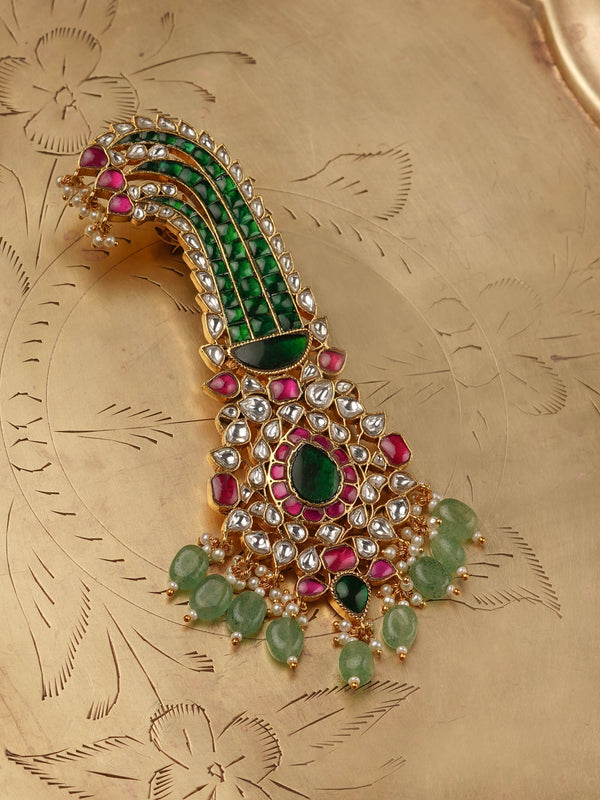 
An ornate gold kalgi (turban ornament) is displayed, featuring intricate green and red stones, kundan work, and pearl drops, set against a textured gold background.