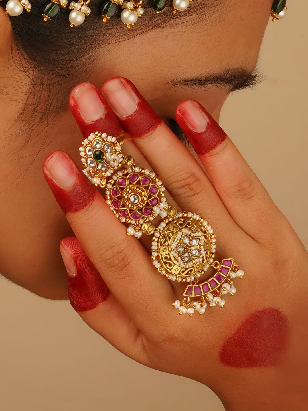 
A hand with fingers dyed red (likely from mehndi or alta) wears an ornate traditional gold ring extending across multiple fingers, adorned with pearls, red and clear stones, and green accents.