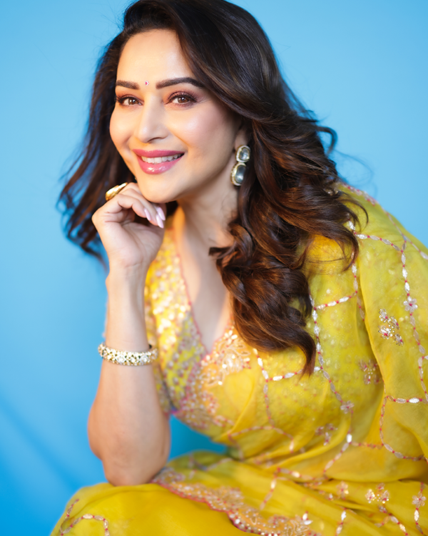 Madhuri Dixit smiles in a yellow outfit, wearing a white and gold bracelet and gold earrings, against a blue background.