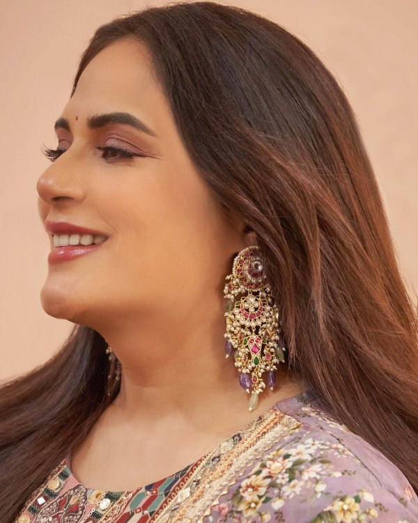 Richa Chadha smiles, wearing a large, ornate multi-colored earring with beads and stones, against a pale pink background.