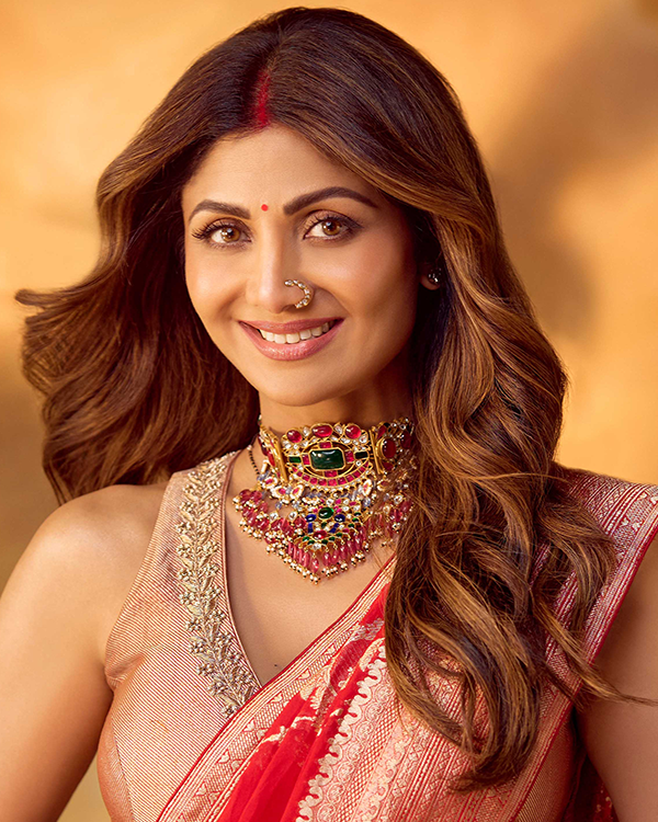 Shilpa Shetty Kundra smiles, adorned with a vibrant choker necklace with green and red stones, a delicate nose ring, and a red bindi and sindoor, wearing a red and gold saree.