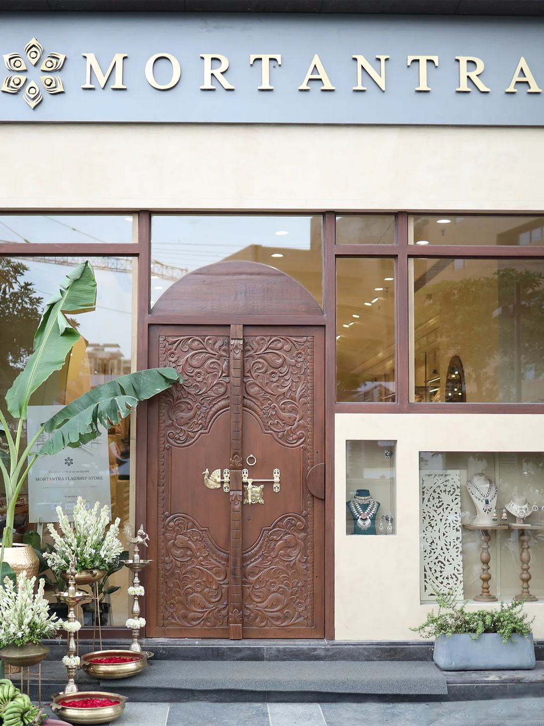 A detailed front view of the Mortantra jewelry store entrance, featuring an ornate carved wooden door, various floral decorations in brass vessels, and display windows showcasing jewelry.