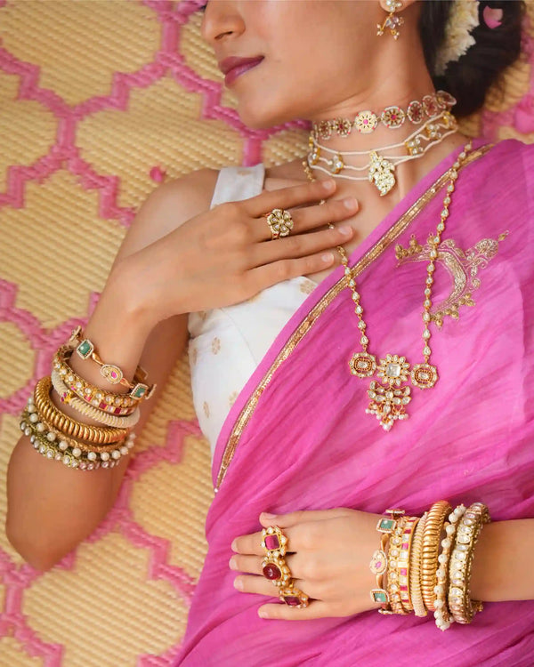 Woman wearing gold jewelry including a necklace, earrings, rings, and bangles on a pink and yellow patterned fabric background.