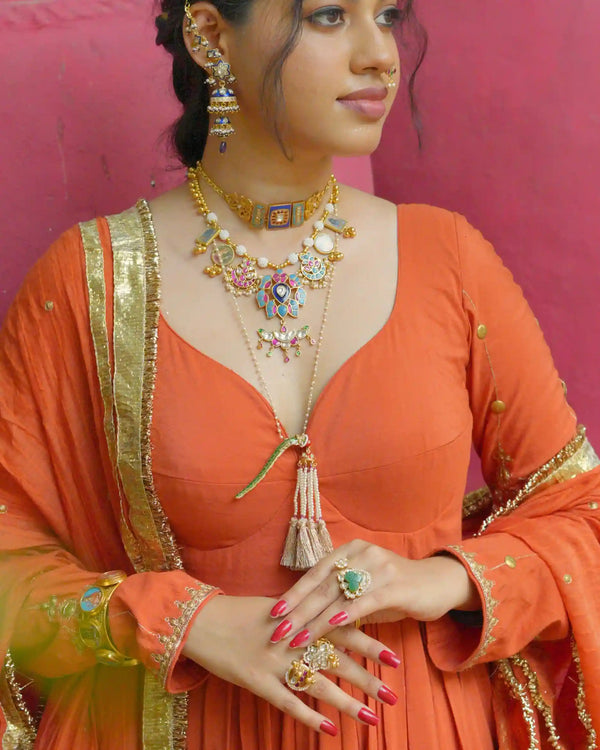 Woman in an orange traditional outfit with gold jewelry against a pink background