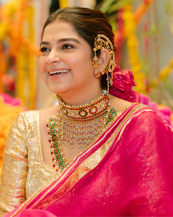 Woman in traditional Indian attire with jewelry, smiling in a decorated setting.