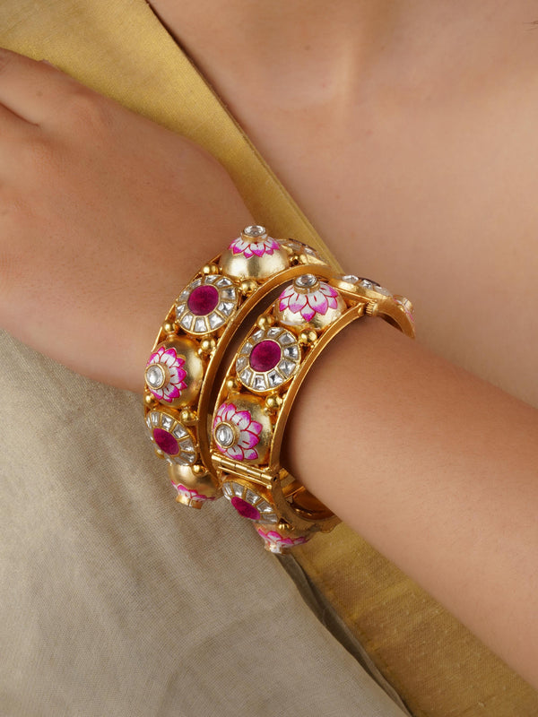 A pair of ornate gold bangles with pink enamel work and ruby-colored stones is worn on a wrist.